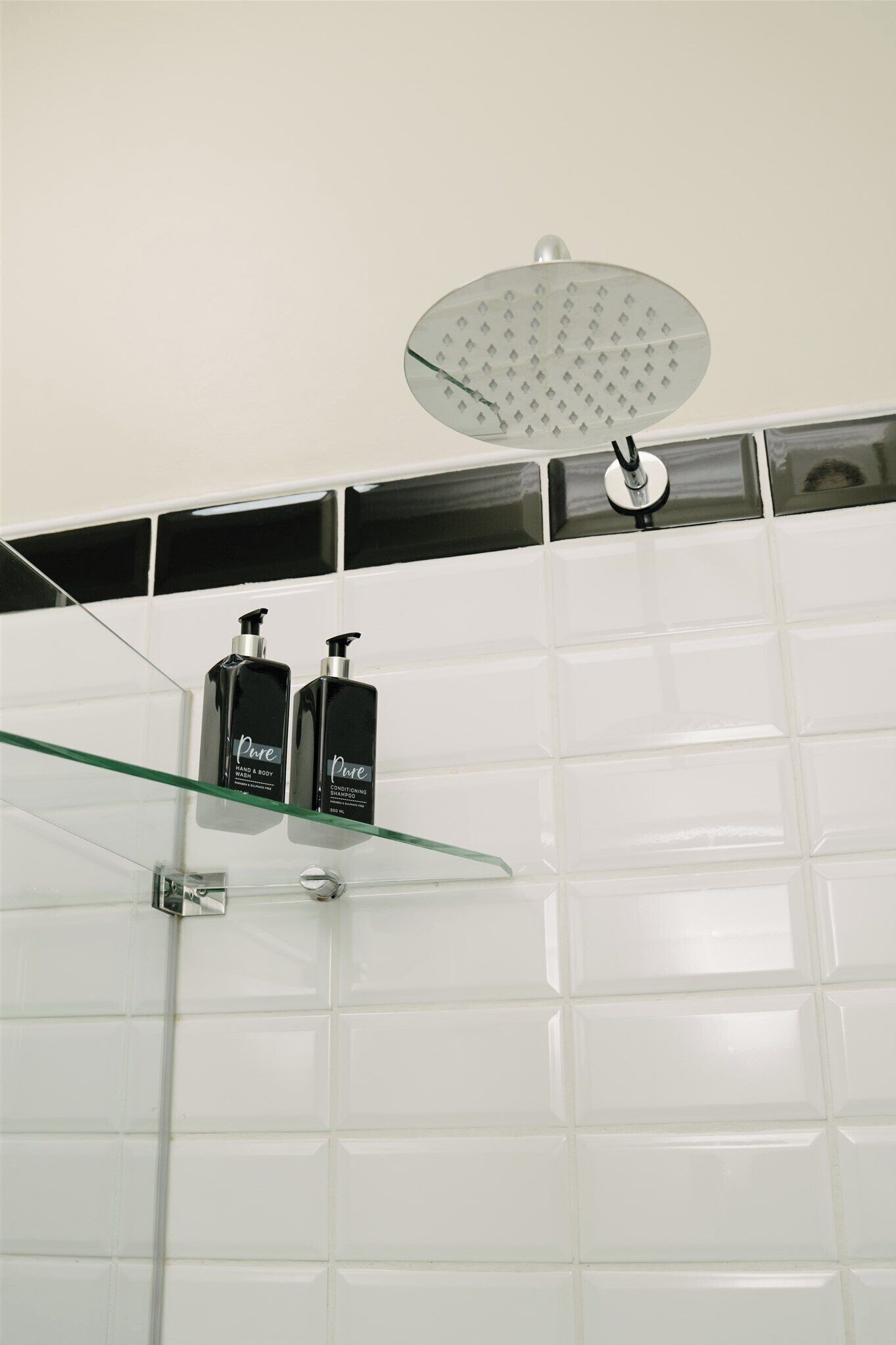 A bathroom with a glass shelf holding two bottles, a showerhead, and white tiled walls.