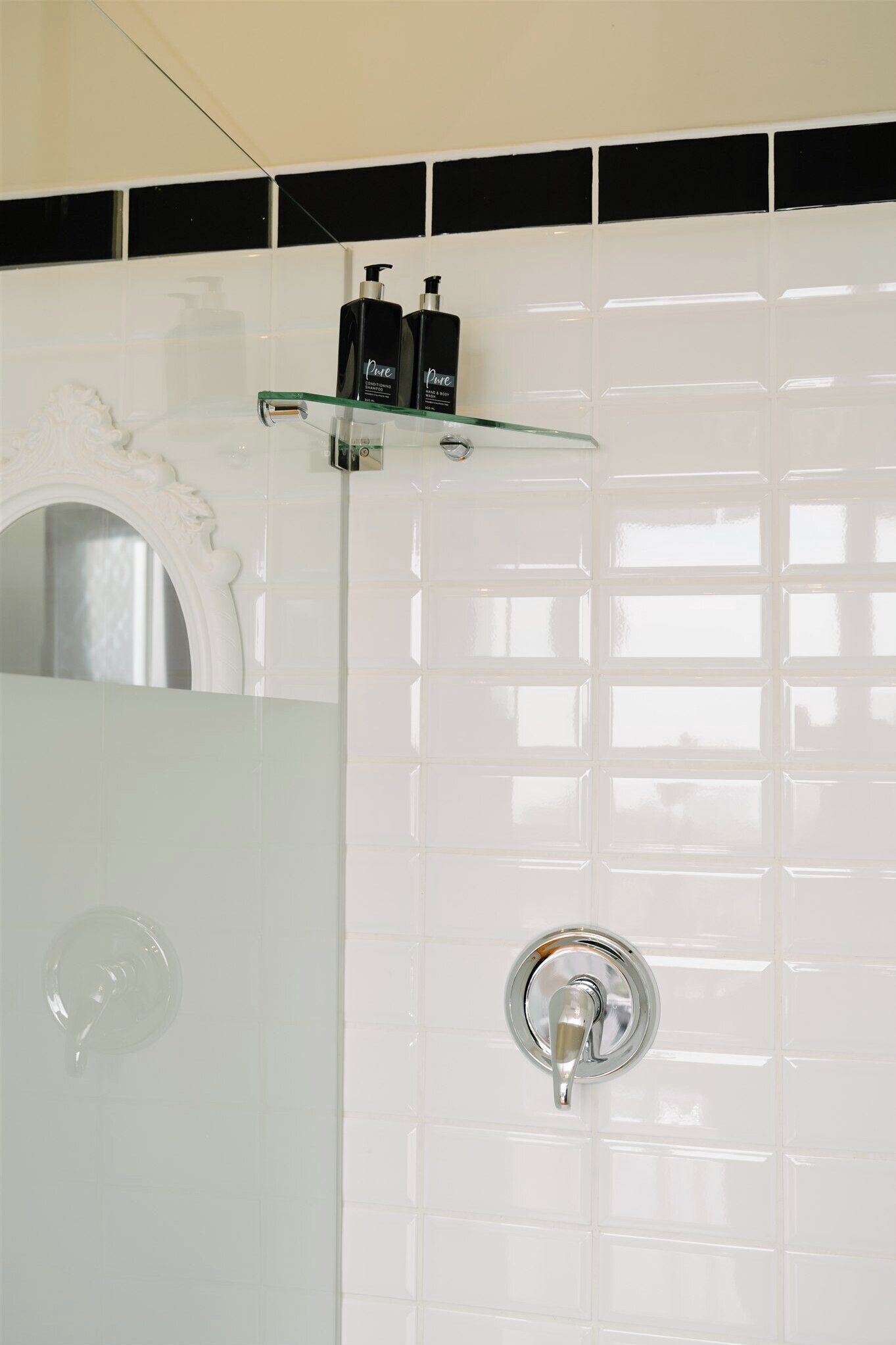 Un baño con paredes de azulejos blancos, cabina de ducha de cristal, lavabo blanco y dos botellas negras sobre una repisa de cristal.
