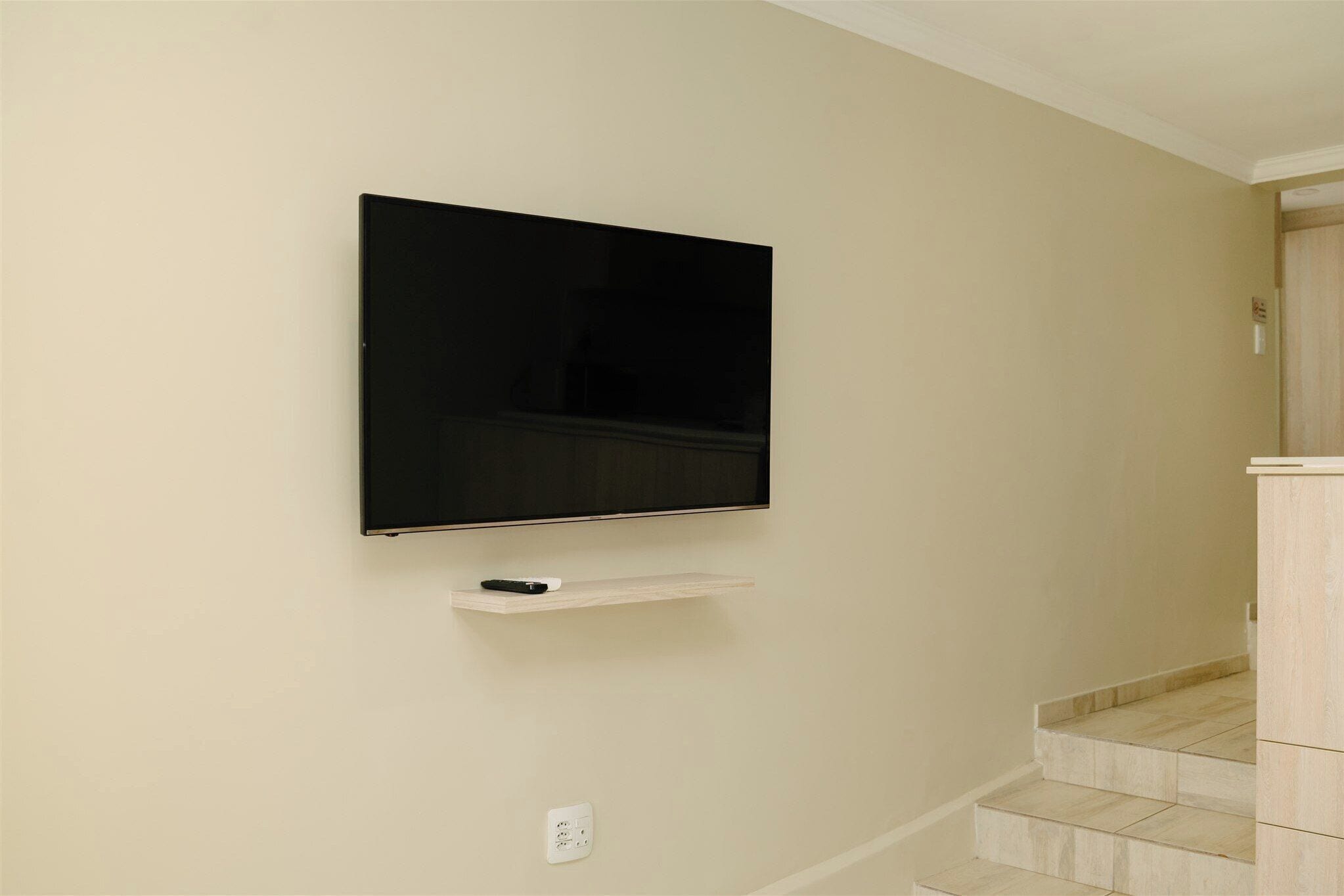 A flat-screen TV mounted on a wall with a remote control on a small shelf below.