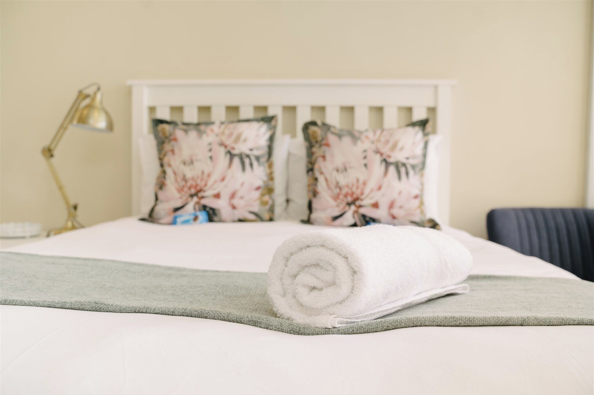 A neatly made bed with a rolled white towel, a green throw blanket, and floral-patterned pillows on a white headboard.
