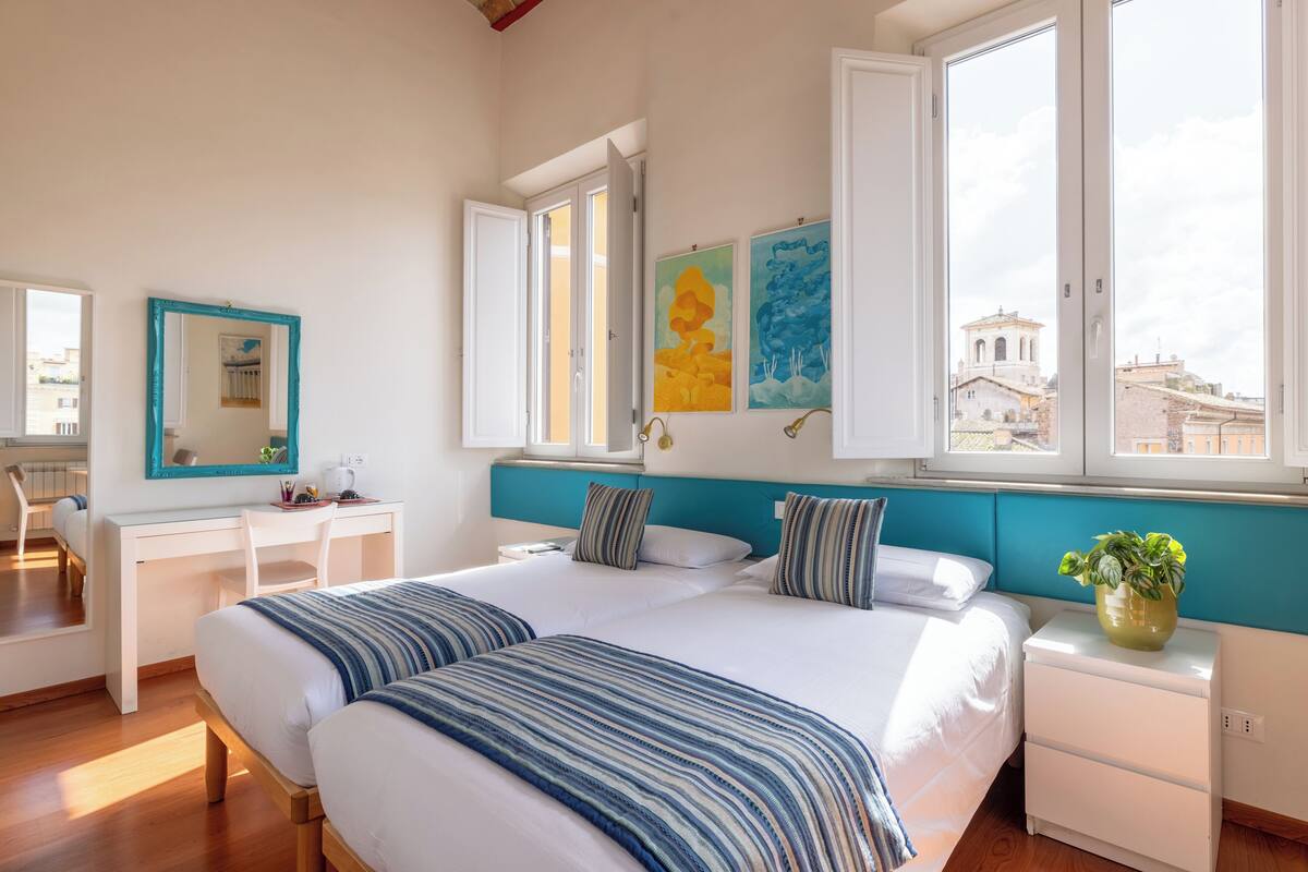 A modern hotel room with a large bed, a desk, and a view of the city through large windows.