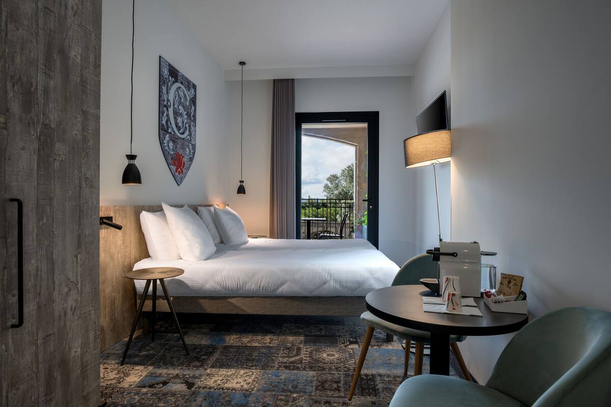 Ein modernes Hotelzimmer mit einem großen Bett, einem kleinen runden Tisch, einem Sessel, einem Schreibtisch und Balkonblick.