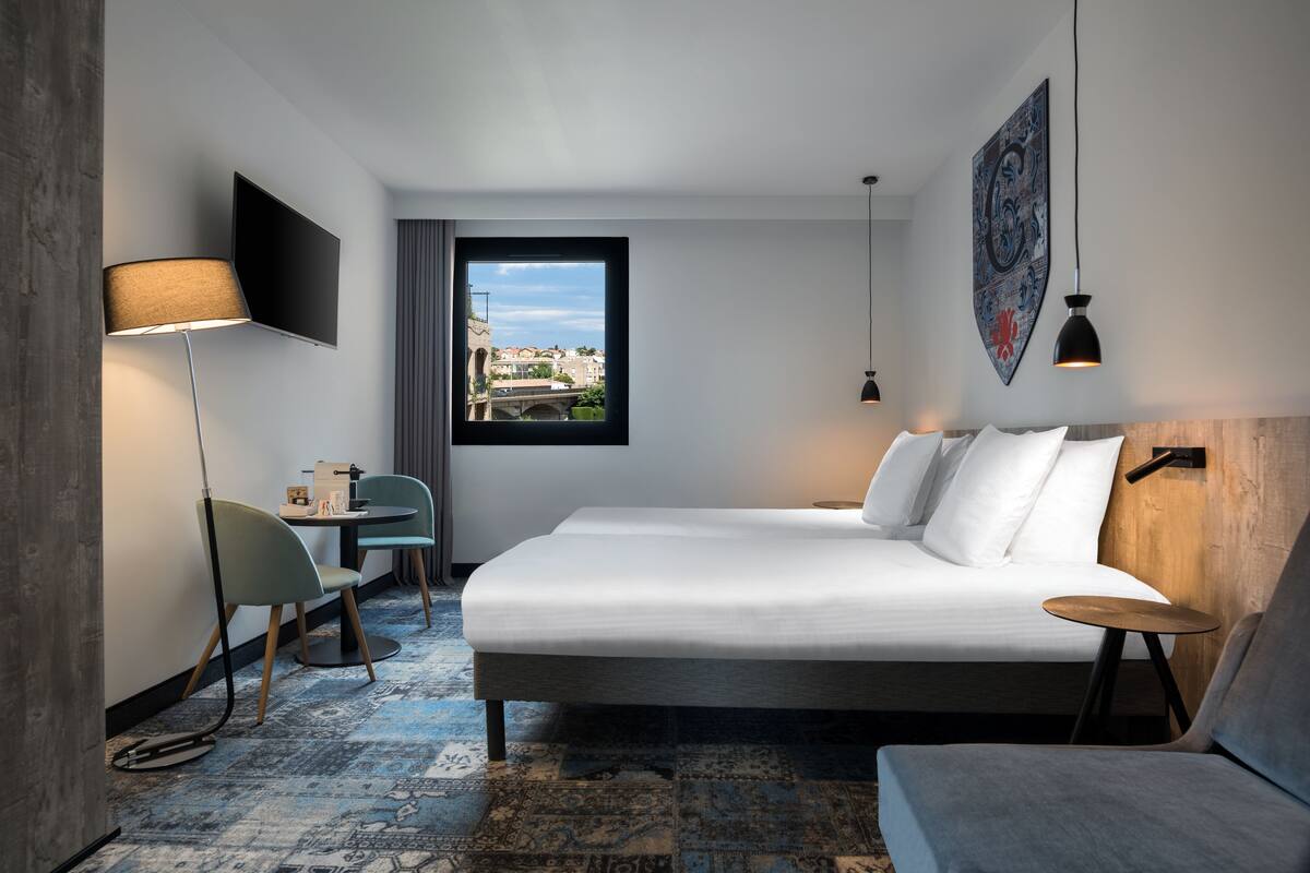 Ein modernes Hotelzimmer mit einem großen Bett, einem Schreibtisch mit Stuhl, einem kleinen Tisch, einer Stehlampe und Blick auf eine Brücke sowie Gebäude durch ein Fenster.