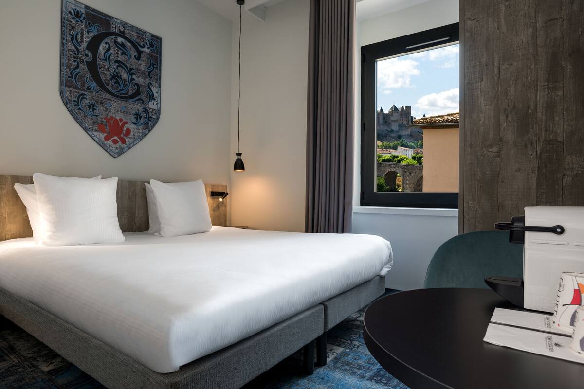 Ein modernes Hotelzimmer mit einem großen Bett, einem Schreibtisch und Blick auf eine Burg durch das Fenster.