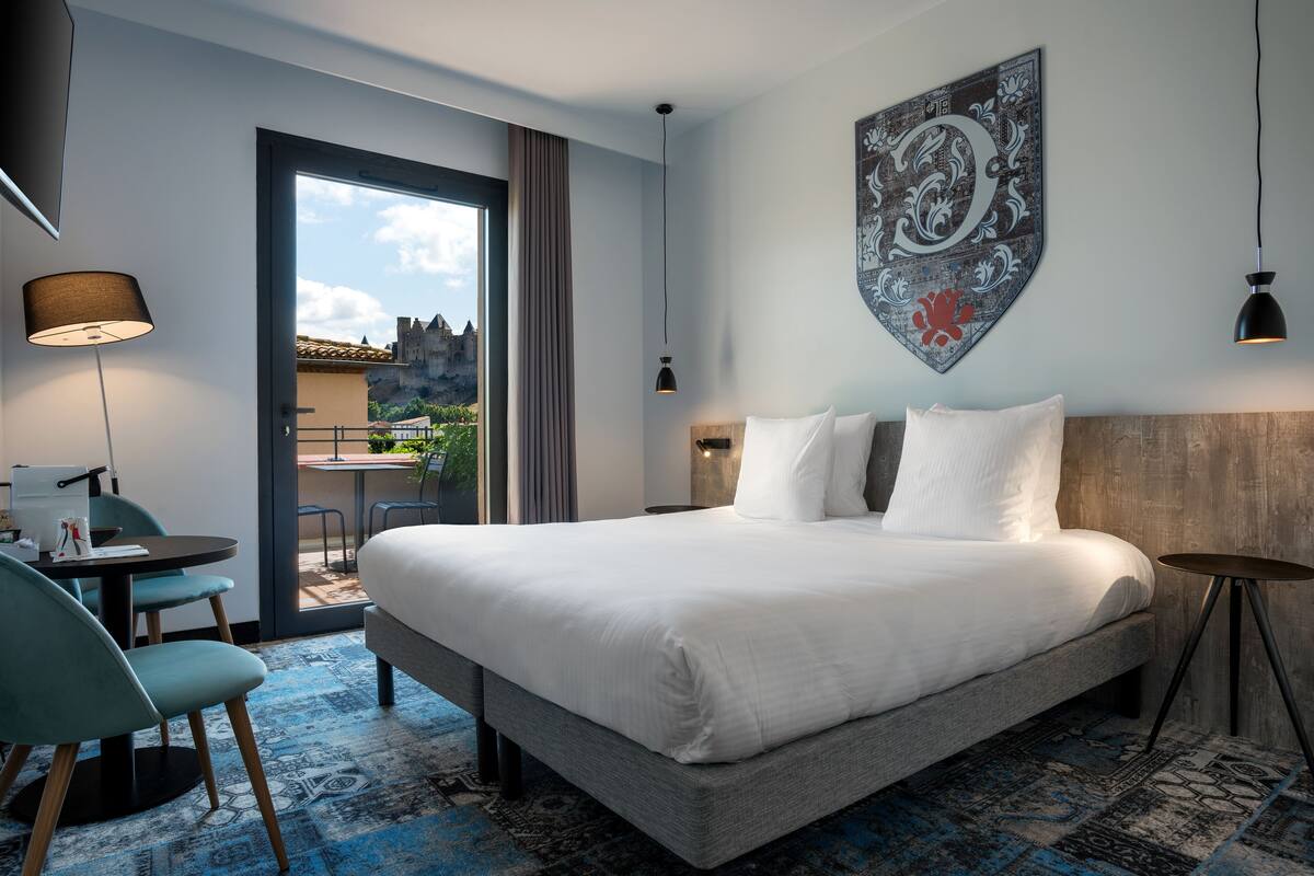 Ein modernes Hotelzimmer mit einem großen Bett, einem Schreibtisch und Blick auf eine Burg durch die Glastür.