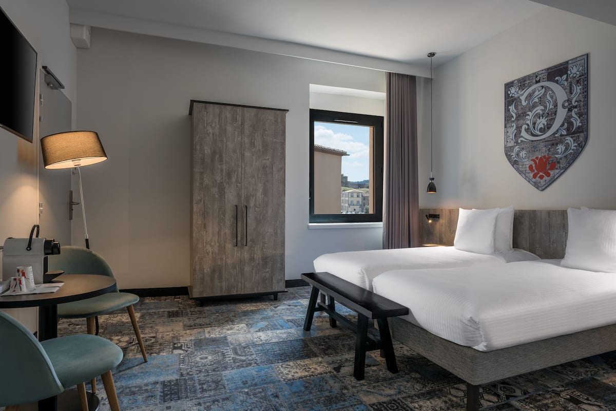 Ein Hotelzimmer mit einem großen Bett, einem Schreibtisch mit Stuhl, einem Kleiderschrank und einem Fenster mit Blick auf Gebäude.
