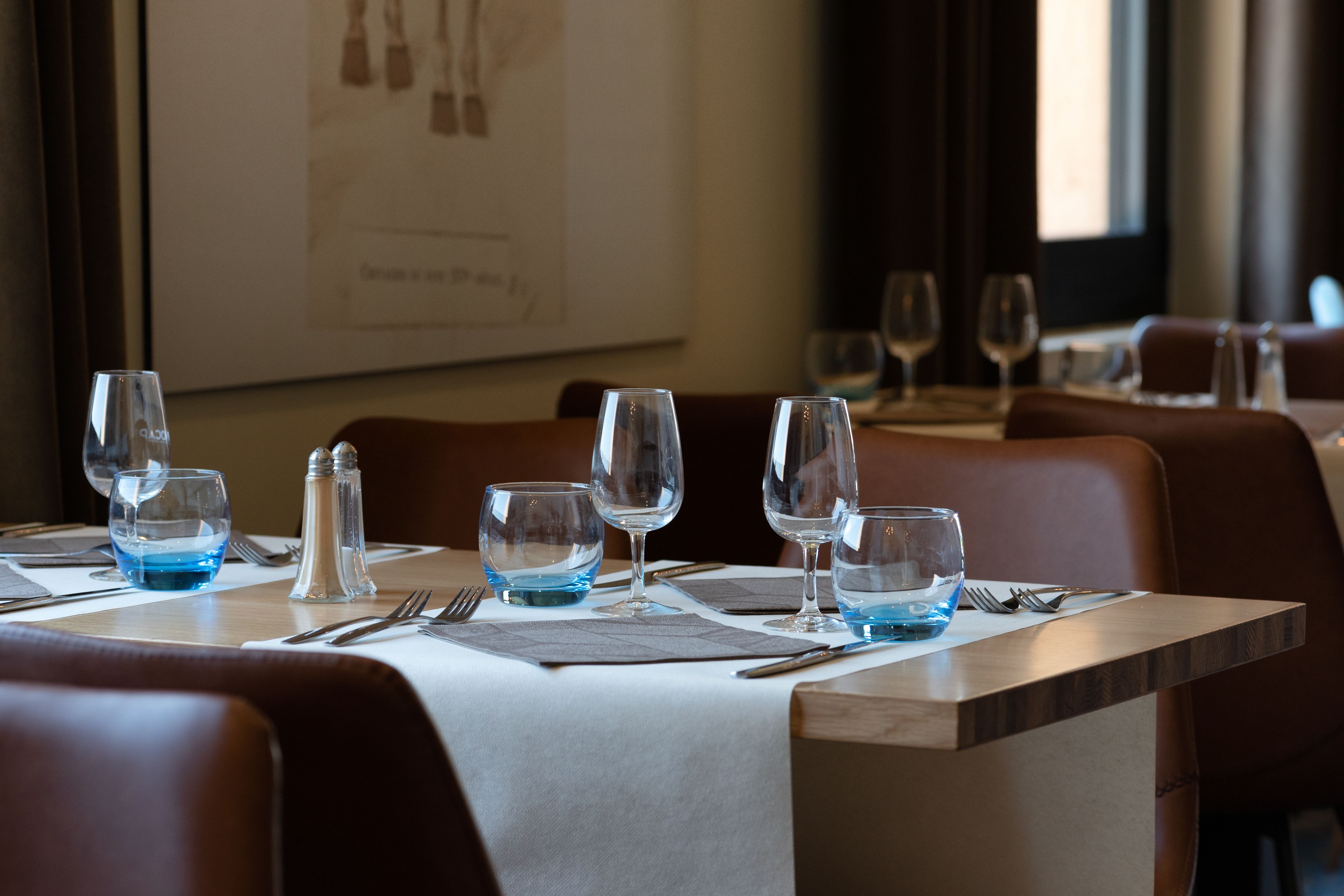 A dining table set with wine glasses, water glasses, and cutlery, with a framed picture on the wall behind.