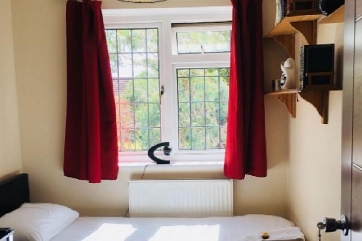 A room with a bed, a window with curtains, a radiator, and a shelf with items.