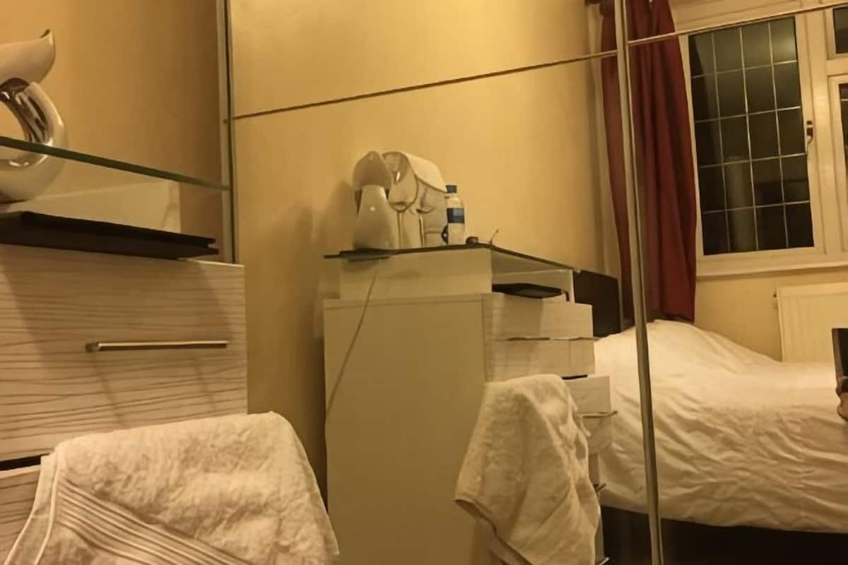 A hotel room with a bed, a bedside table, a mirror, a window with curtains, and a small refrigerator.