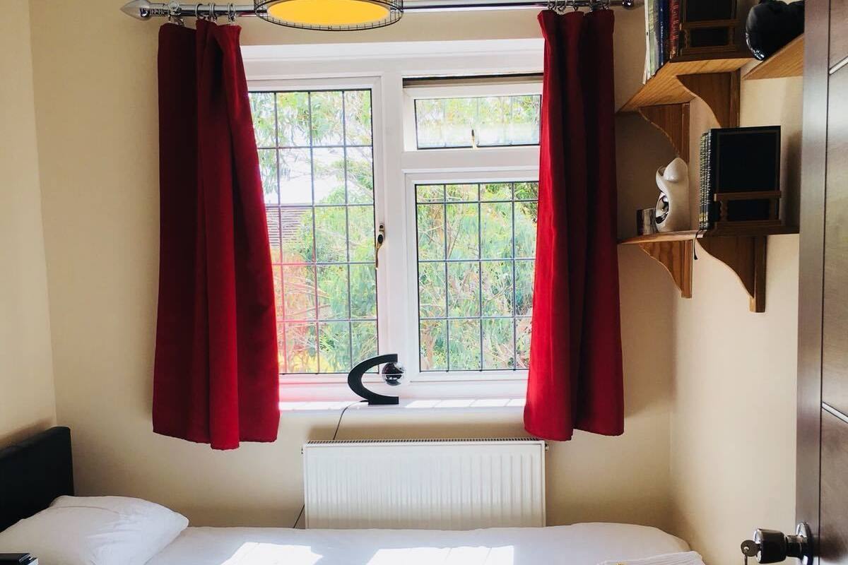A bedroom with a bed, a radiator, a window with curtains, a hanging light fixture, and a small shelf with books and a decorative item.