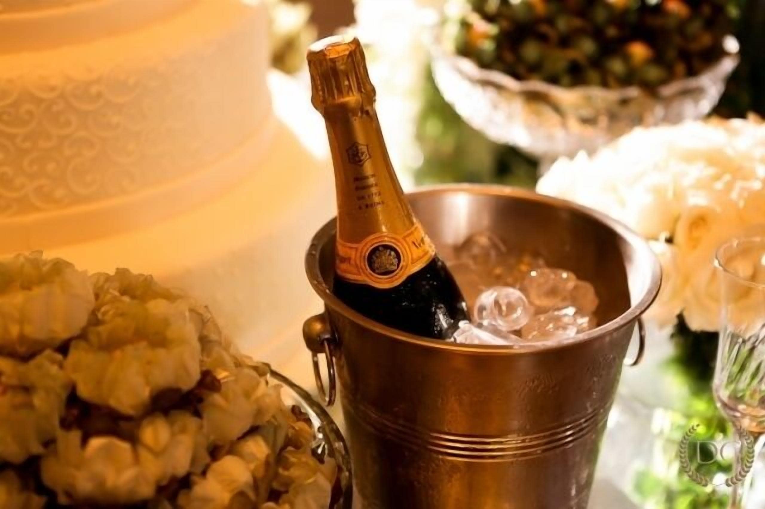 A champagne bottle in a metal bucket with ice, surrounded by floral arrangements and a cake.
