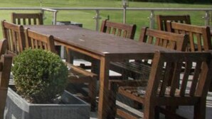 An outdoor seating area with wooden tables and chairs, a potted plant, and a view of a weeping willow tree. - Bianca's House Hotel Heathrow Airport (Slough)
