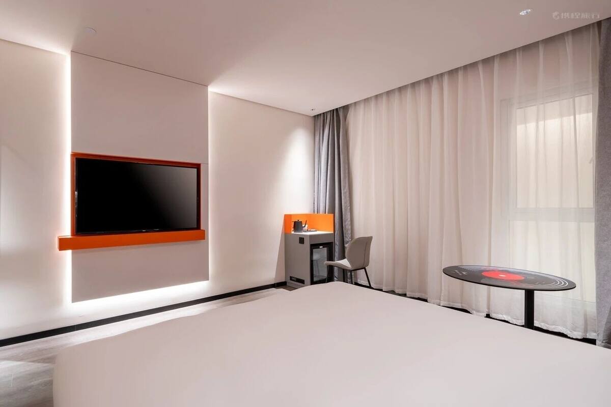 A modern hotel room with a large bed, a flat-screen TV mounted on the wall, a small desk with a chair, and a round table with a red object on it.