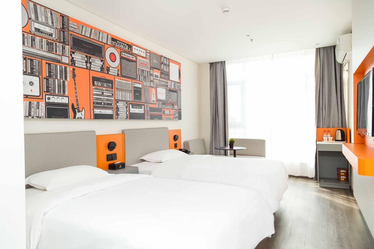 A hotel room with two beds, a wall decorated with musical equipment, a desk with a chair, and a window with curtains.