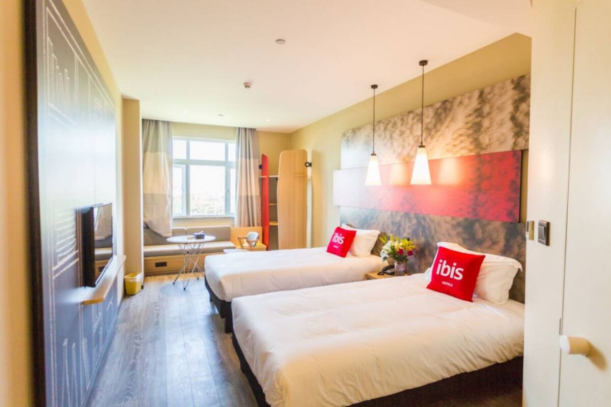 Quarto de hotel com duas camas, uma escrivaninha e uma janela grande.
