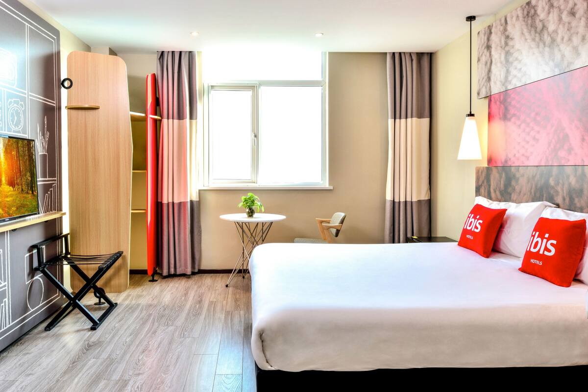 A hotel room with a bed, a small table, a chair, a wardrobe, and a window.