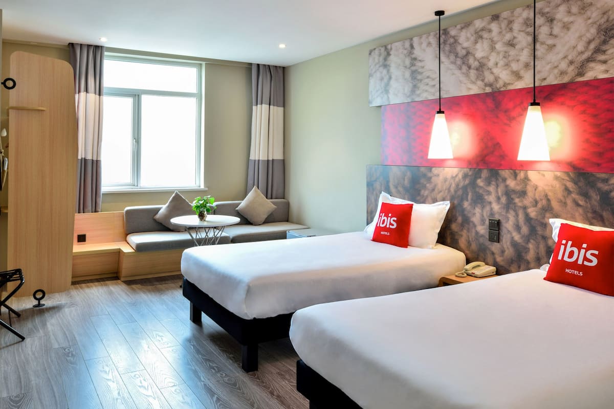 A hotel room with two beds, a sofa, a small table with a plant, and a window with curtains.