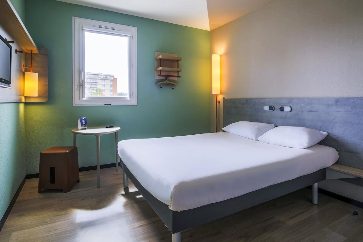 Une chambre d’hôtel comprenant un lit, une petite table, un tabouret, un téléviseur fixé au mur, une fenêtre et une lampe fixée au mur.