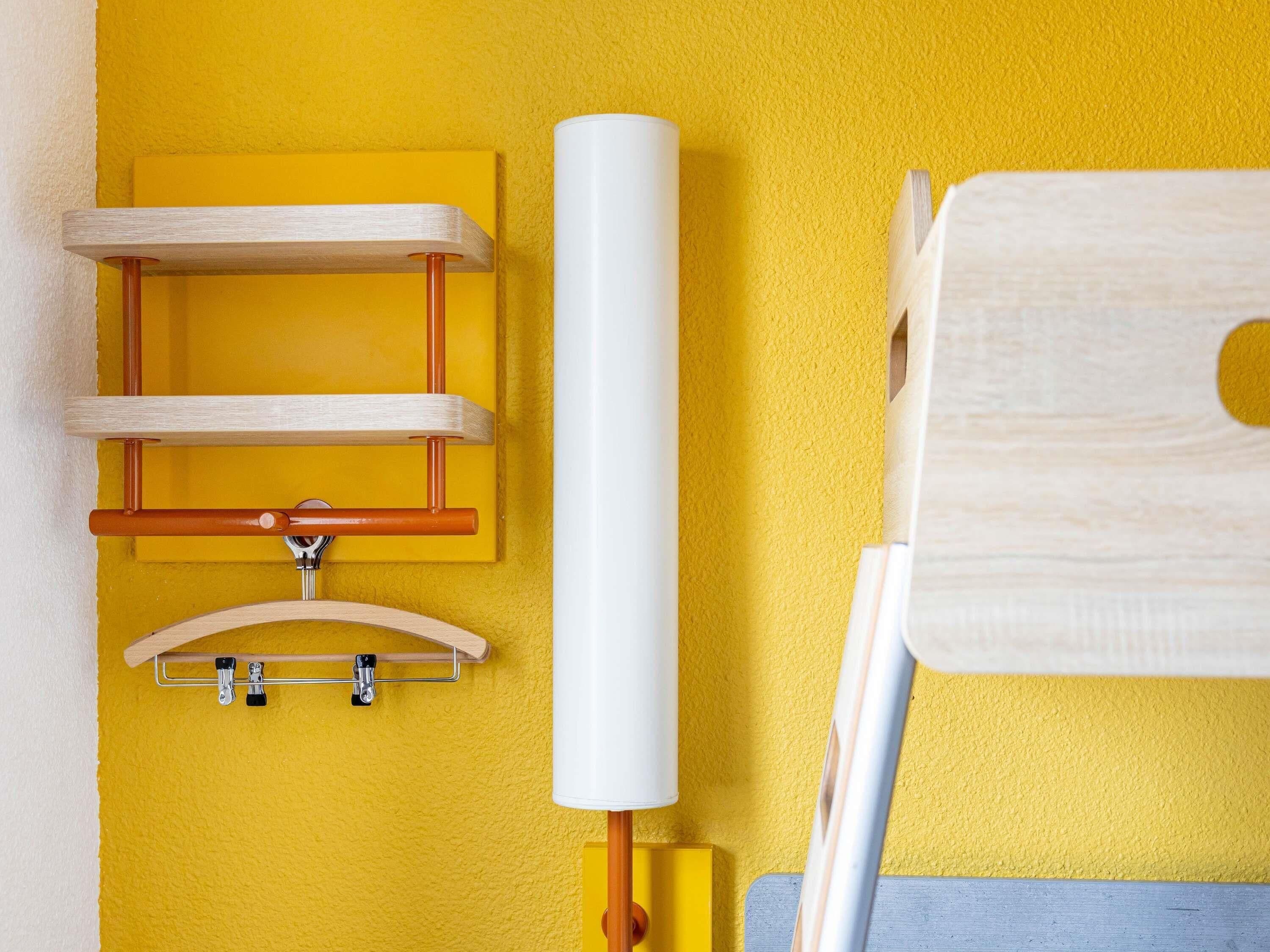 A bathroom with a yellow wall, a wooden shelf, a white cylindrical object, and a wooden chair.