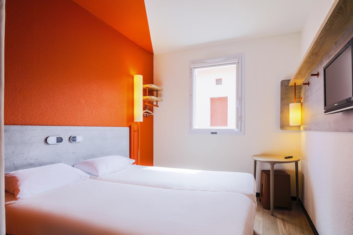 Une chambre d’hôtel avec un lit, des commandes situées près du lit, une petite table, une télévision et une fenêtre.