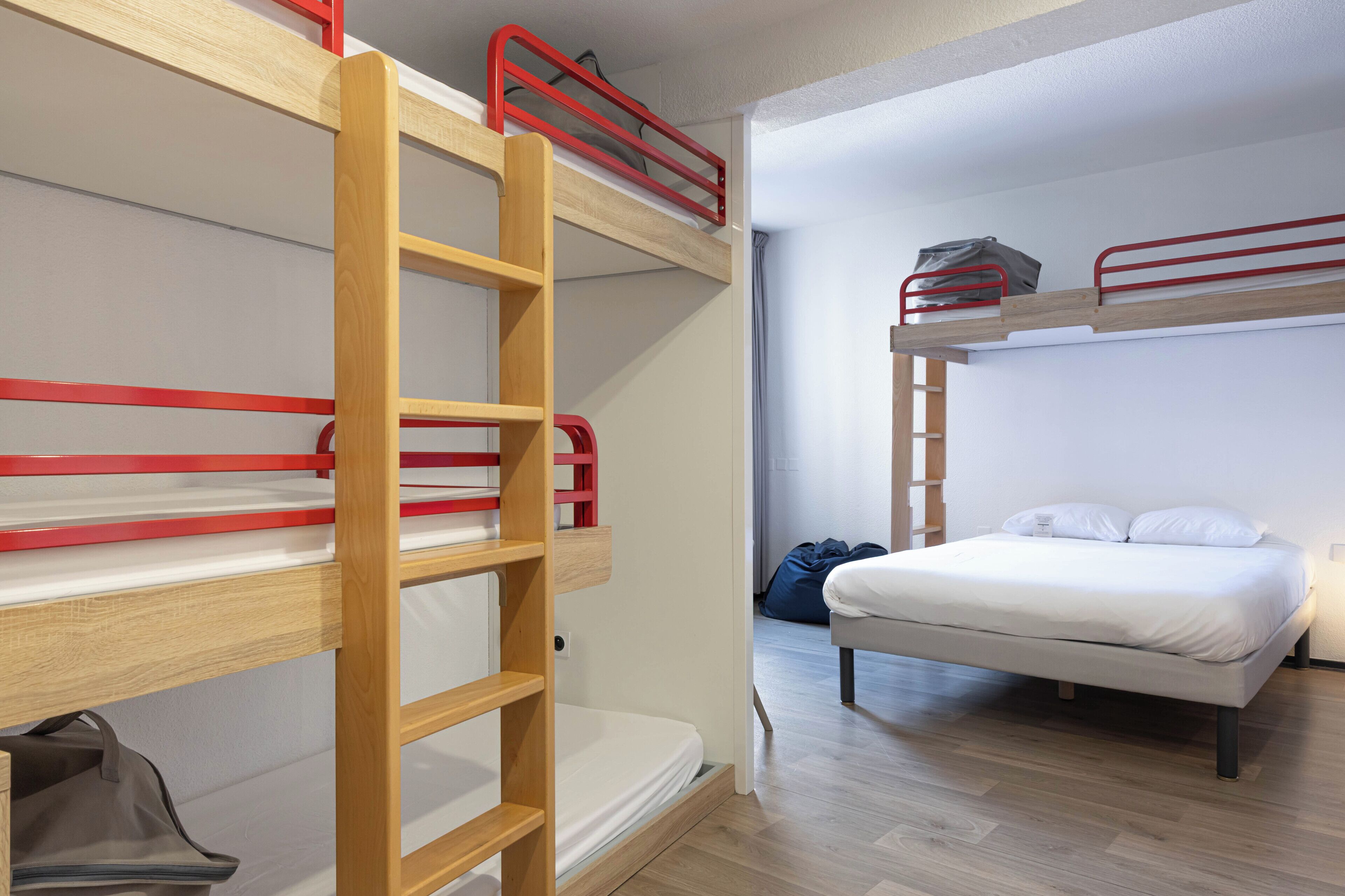 Une chambre avec un lit superposé, une échelle en bois, un lit simple et un sac posé sur le sol.