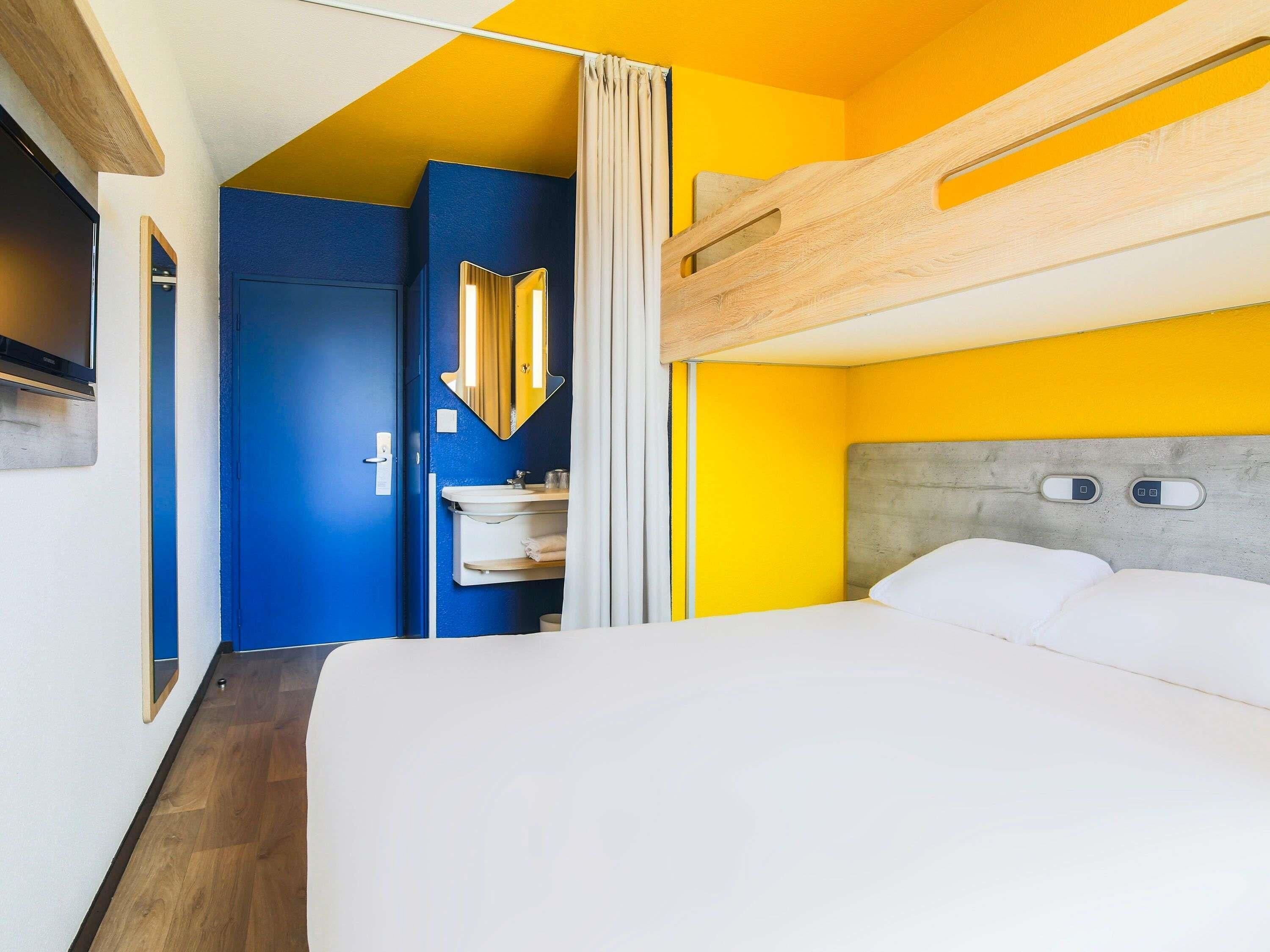 Une chambre d’hôtel avec un lit superposé, une salle de bain avec un miroir et un lavabo, et une porte donnant sur une autre chambre.