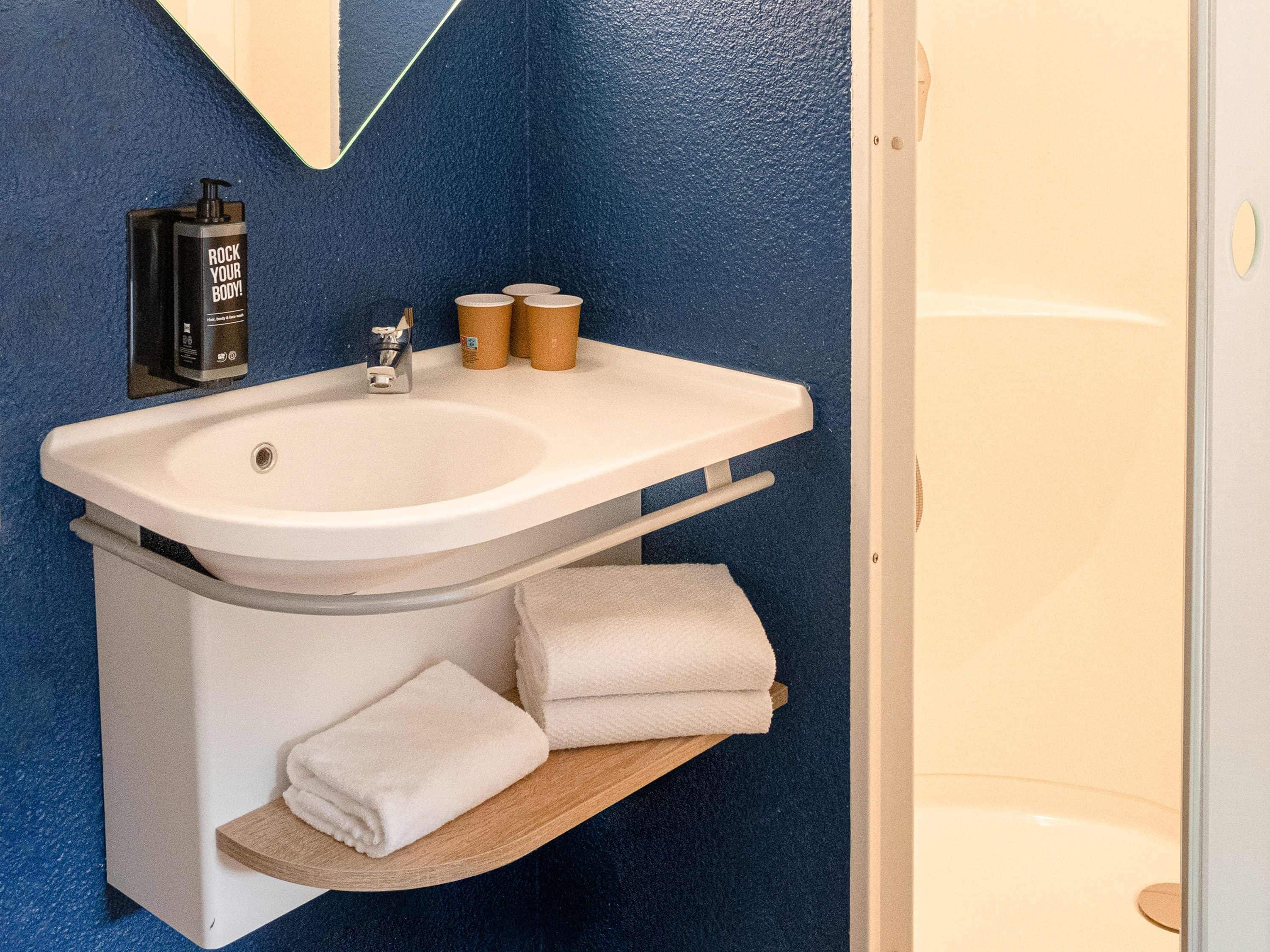 Un baño con lavamanos blanco, un espejo, toallero y una botella de champú.