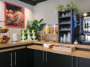 Une cuisine Ă©quipĂ©e dâune machine Ă cafĂ©, dâun panier de fruits, de boĂźtes de conservation vertes, dâun distributeur de jus et dâune autre machine Ă cafĂ©.