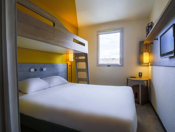 Une chambre d’hôtel compacte comprenant un lit superposé, une petite table de chevet, un téléviseur fixé au mur et une fenêtre laissant entrer la lumière naturelle.