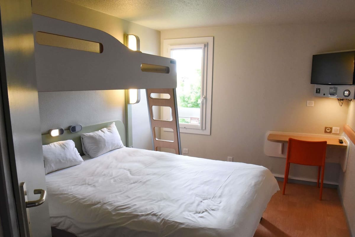 Quarto de hotel com cama de solteiro, escrivaninha com cadeira, televisão fixada na parede e janela que ilumina naturalmente.