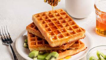 A stack of waffles with honey being drizzled on top, accompanied by sliced kiwi fruit.