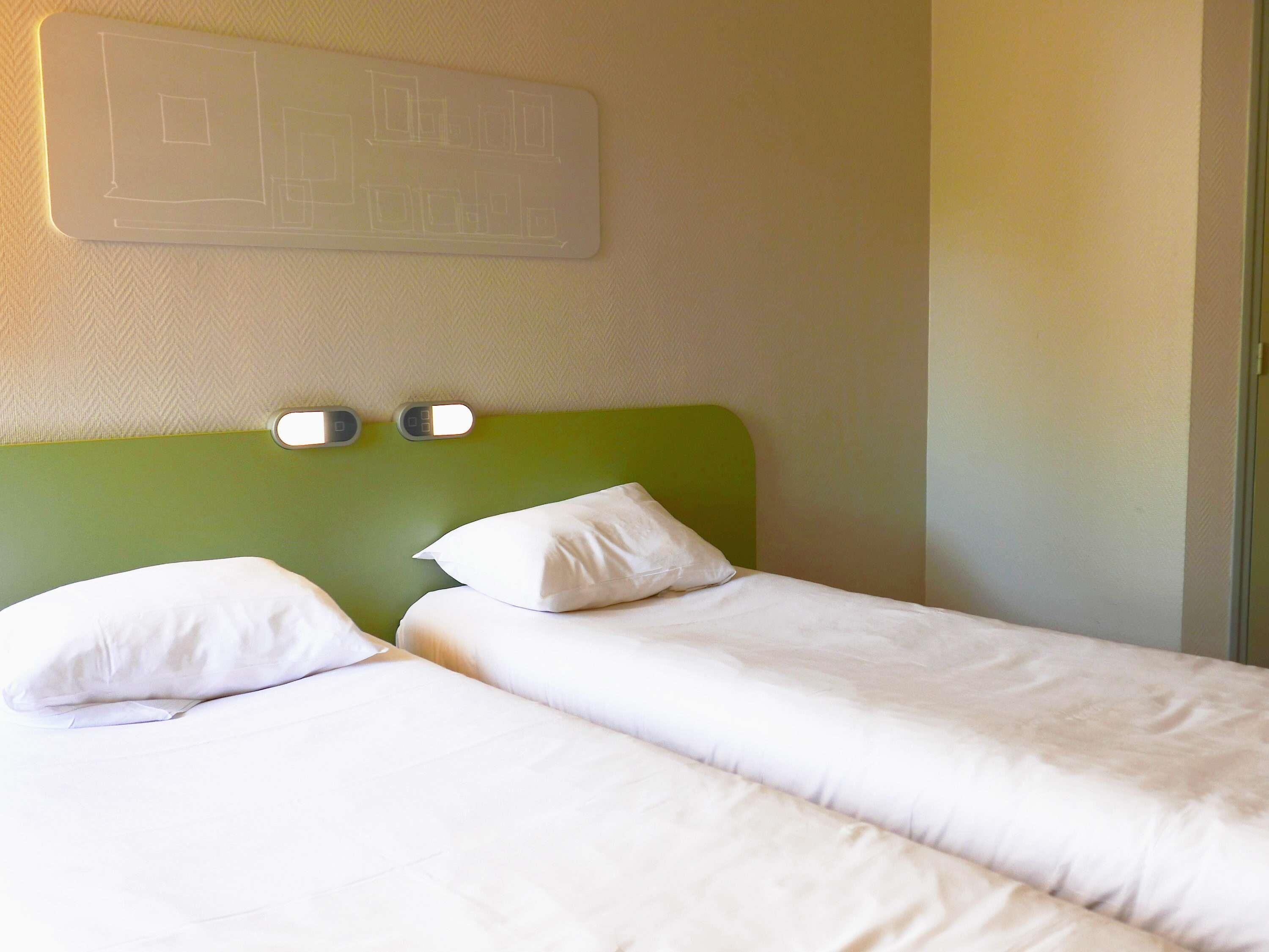 A hotel room with two single beds, a green headboard, and a wall-mounted light switch.