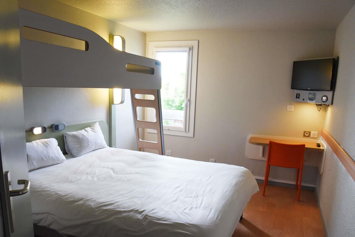 A single bed with a headboard, a desk with a chair, a television mounted on the wall, and a window with a view of greenery.