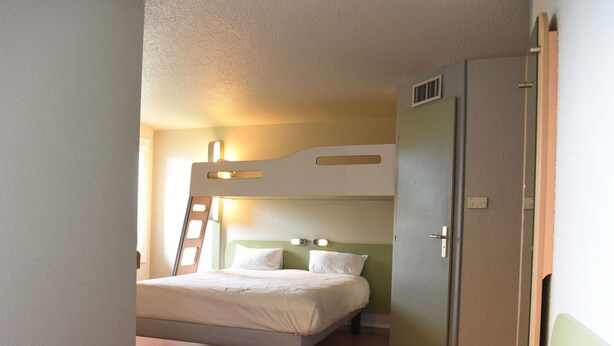 Un lit superposé avec une échelle, un lit simple et une armoire dans une pièce dont le sol et les murs sont de couleur claire.