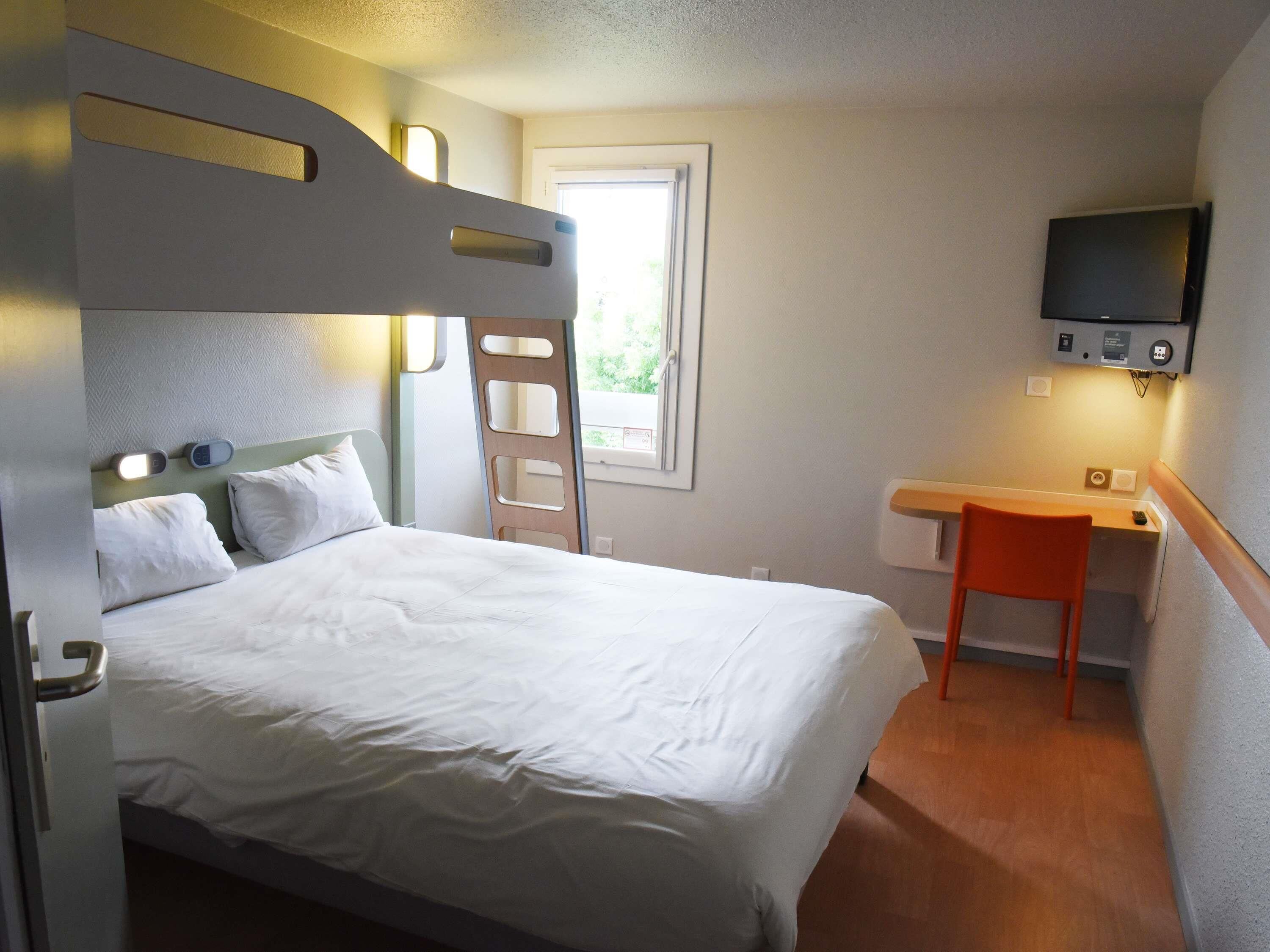 A single bed with a headboard, a desk with a chair, a television mounted on the wall, and a window with a view of greenery.