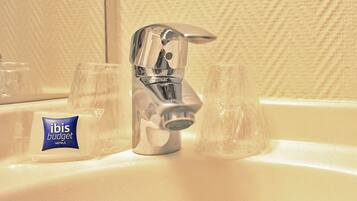Un lavabo de salle de bain avec un robinet chromé, deux verres transparents et un petit ensemble de produits de toilette de l’hôtel ibis budget.