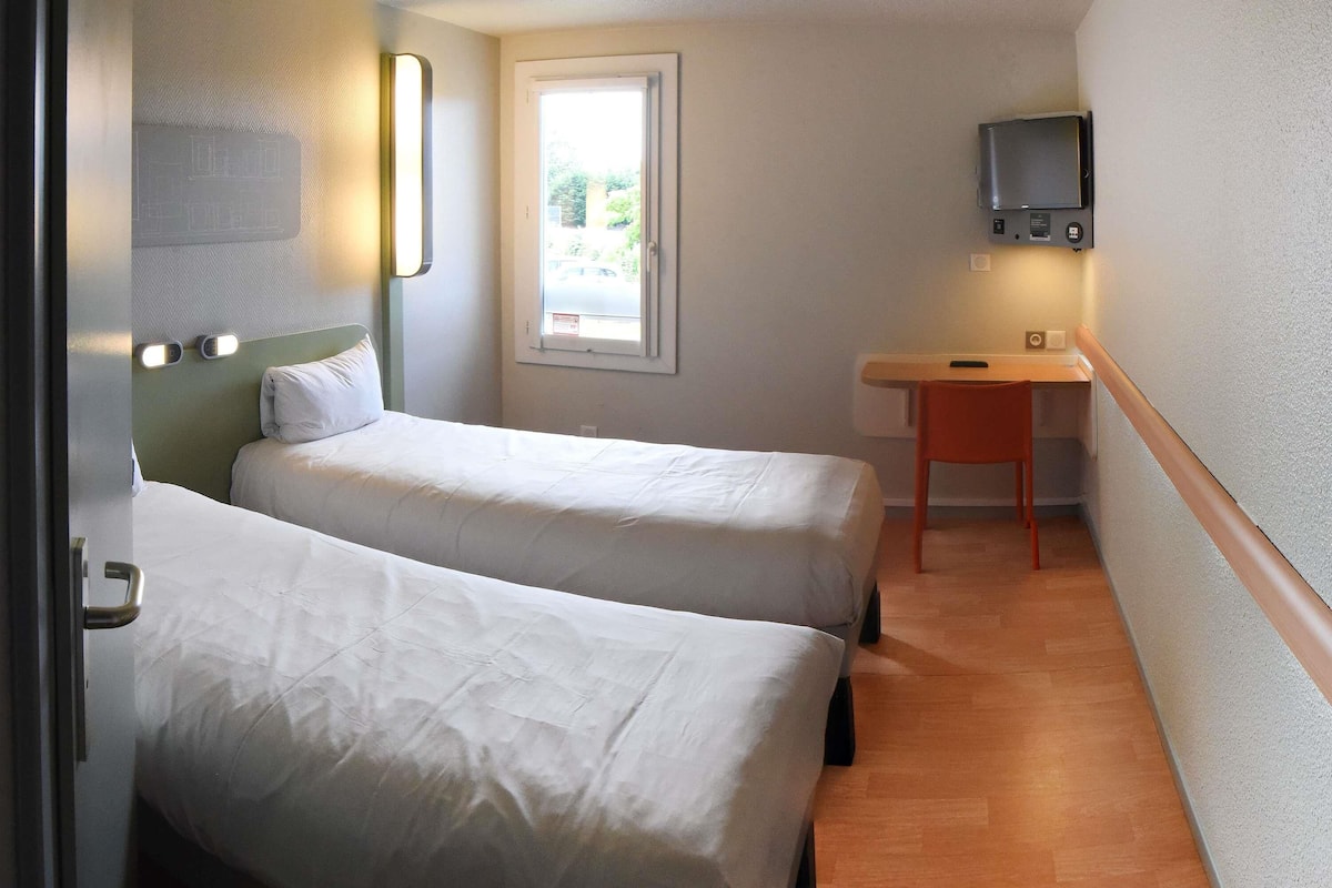 Quarto pequeno e compacto com duas camas individuais, uma escrivaninha, uma cadeira, uma televisão e uma janela com vista para o exterior.