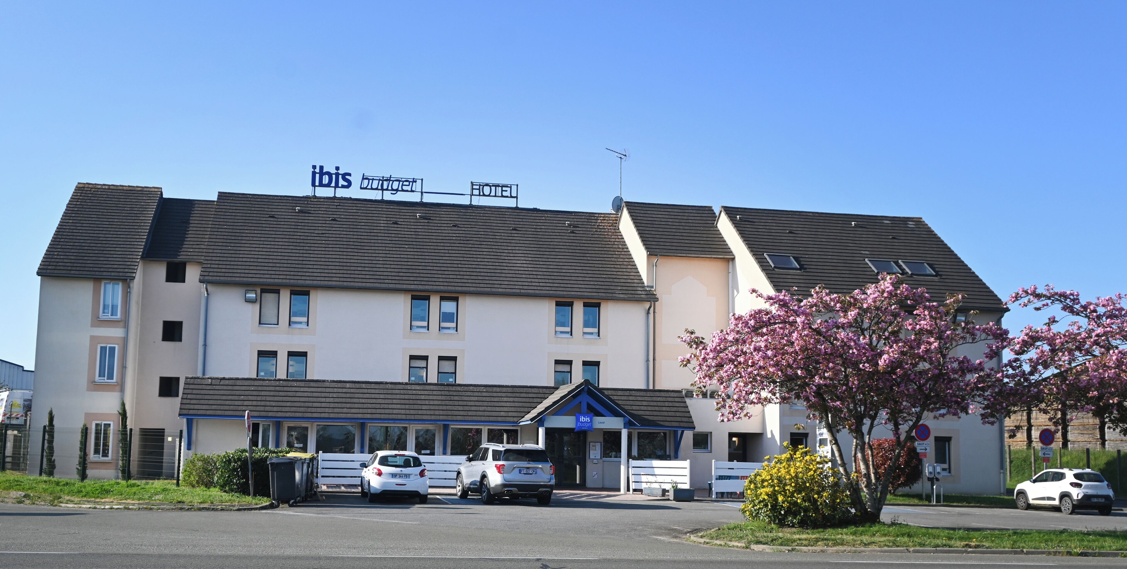 Et hotel med et skilt, der viser 'ibis budget hotel' og 'HOTEL'. Der er parkerede biler foran, en lille have med lyserøde blomster og en klar blå himmel.