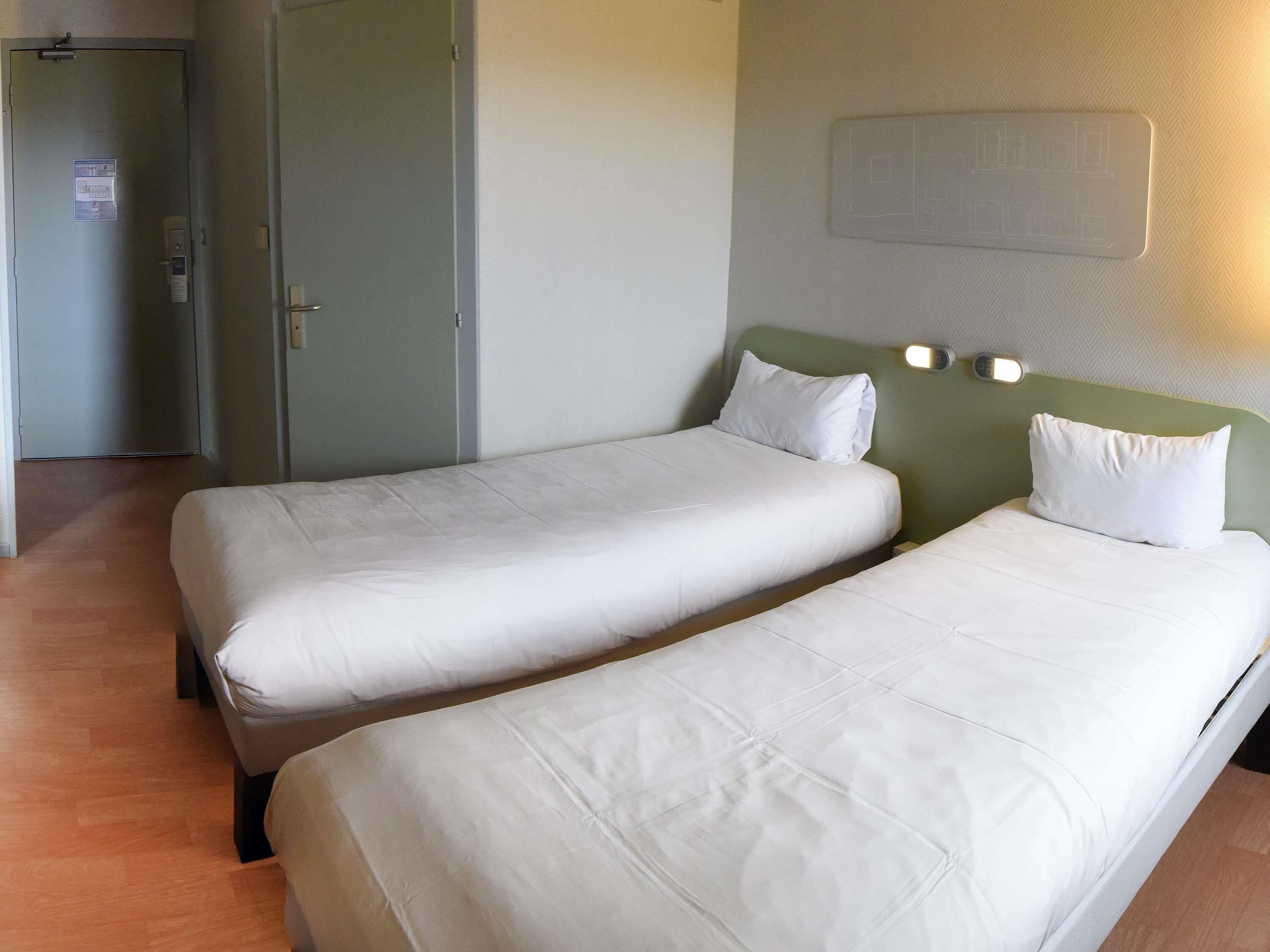 Une chambre d’hôtel avec deux lits simples, une porte à serrure et un luminaire fixé au mur.