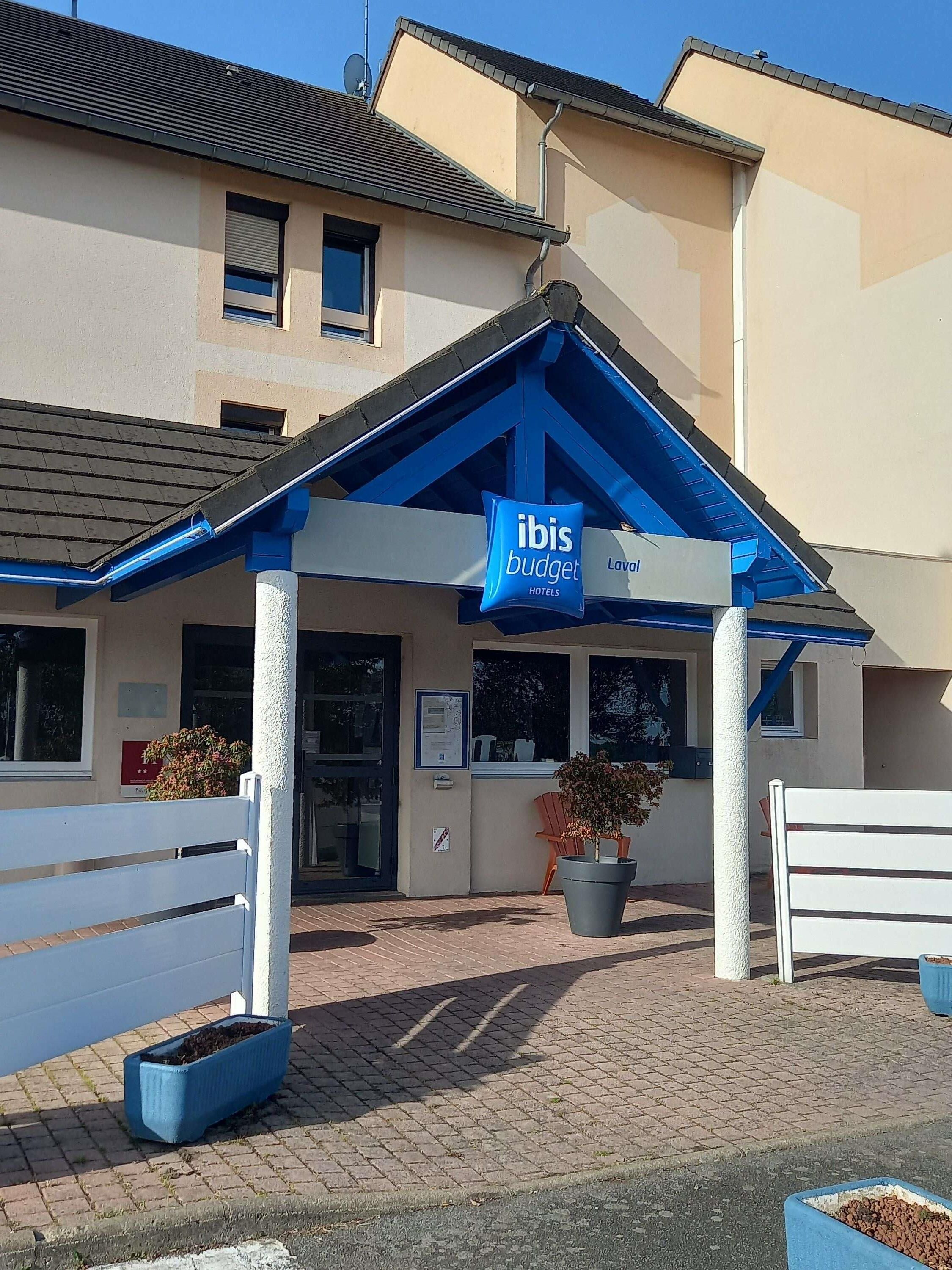 Een hotelingang met een blauw luifel, een bord voor ibis budget hotels en een verhard gebied met bankjes en plantenbakken.