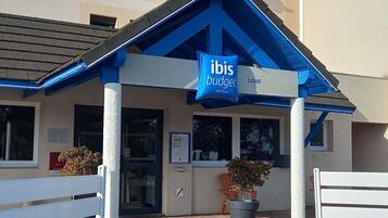 Een hotelingang met een blauw luifel, een bord voor ibis budget hotels en een verhard gebied met bankjes en plantenbakken.