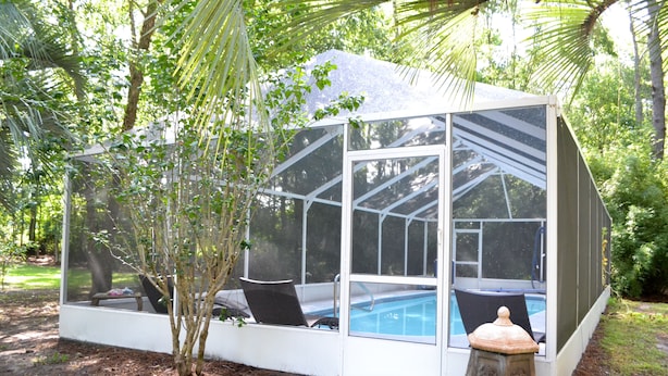 Una casa de piscina con paredes de vidrio, con un banco y una pequeña mesa, rodeada de árboles y un jardín.
