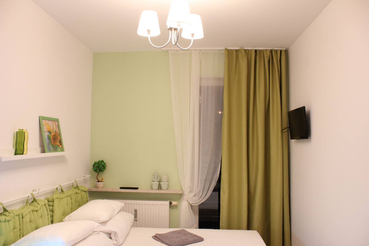 Une chambre à coucher comprenant un lit, une table de chevet, un téléviseur fixé au mur et une fenêtre avec des rideaux.
