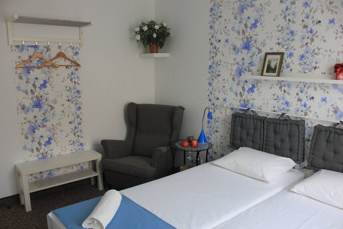 Une chambre d’hôtel avec un lit, une chaise, une petite table, un banc et un mur recouvert d’un papier peint à motifs floraux.