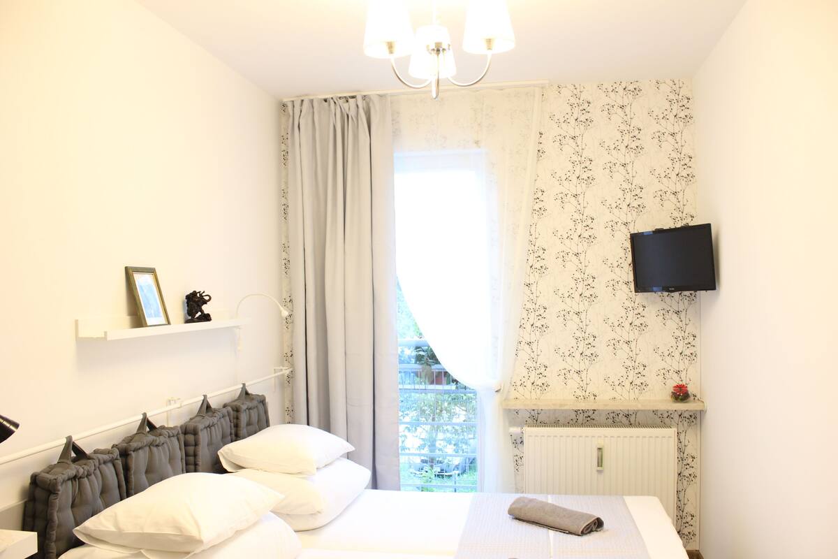 Une chambre à coucher avec un lit, un téléviseur fixé au mur, une fenêtre avec des rideaux et un radiateur.