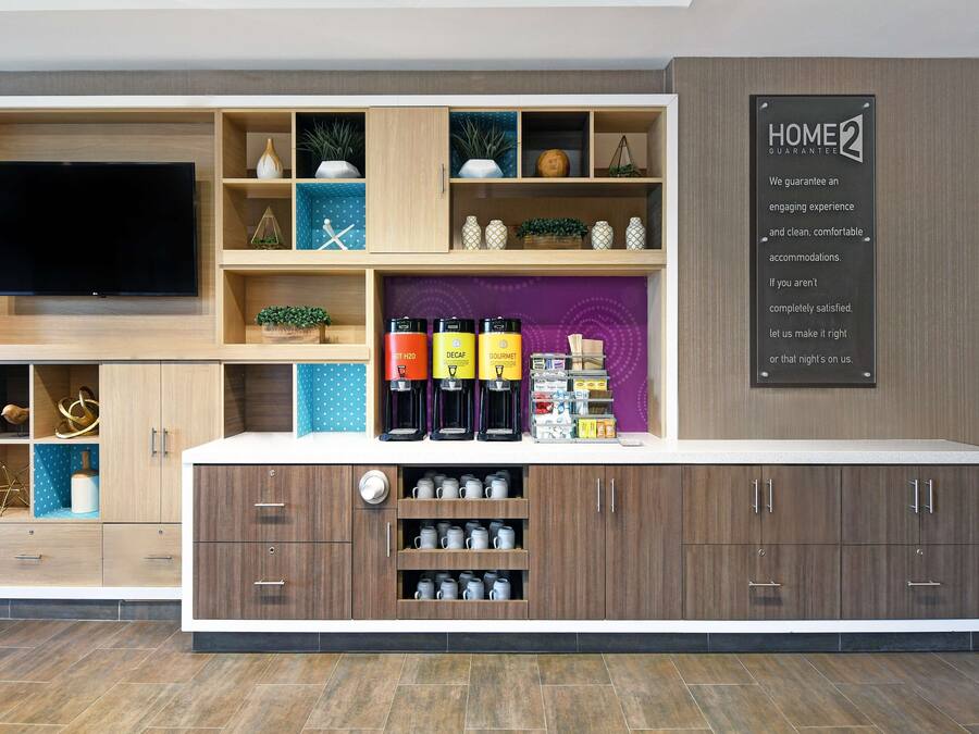 A modern hotel reception area with a flat-screen TV, a coffee station, and a sign stating "HOME 2 GUARANTEE".