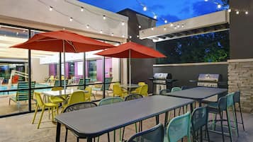 An outdoor dining area with tables and chairs, string lights, and a pool in the background.