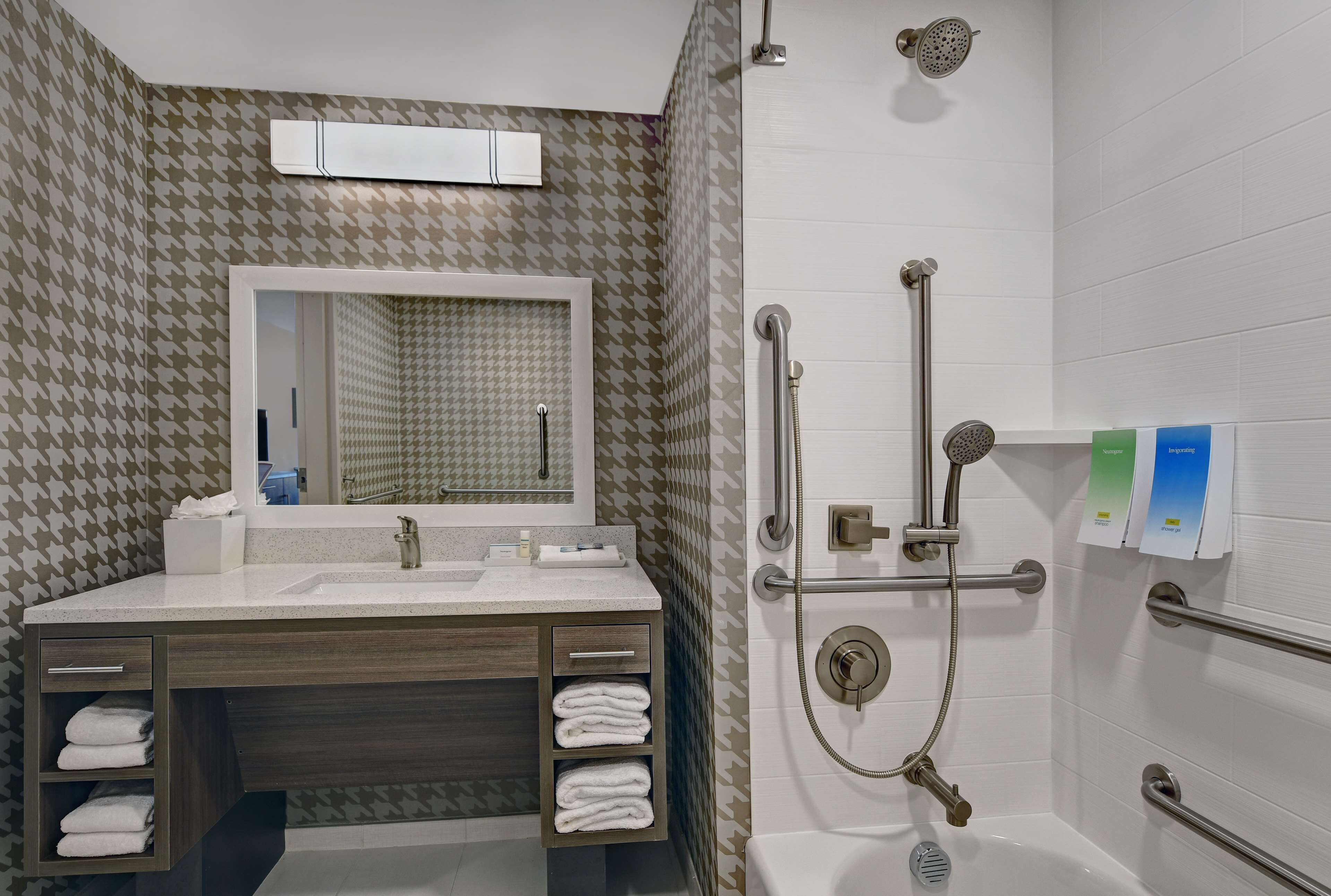 A modern bathroom with a vanity, mirror, towel racks, and a walk-in shower with grab bars.