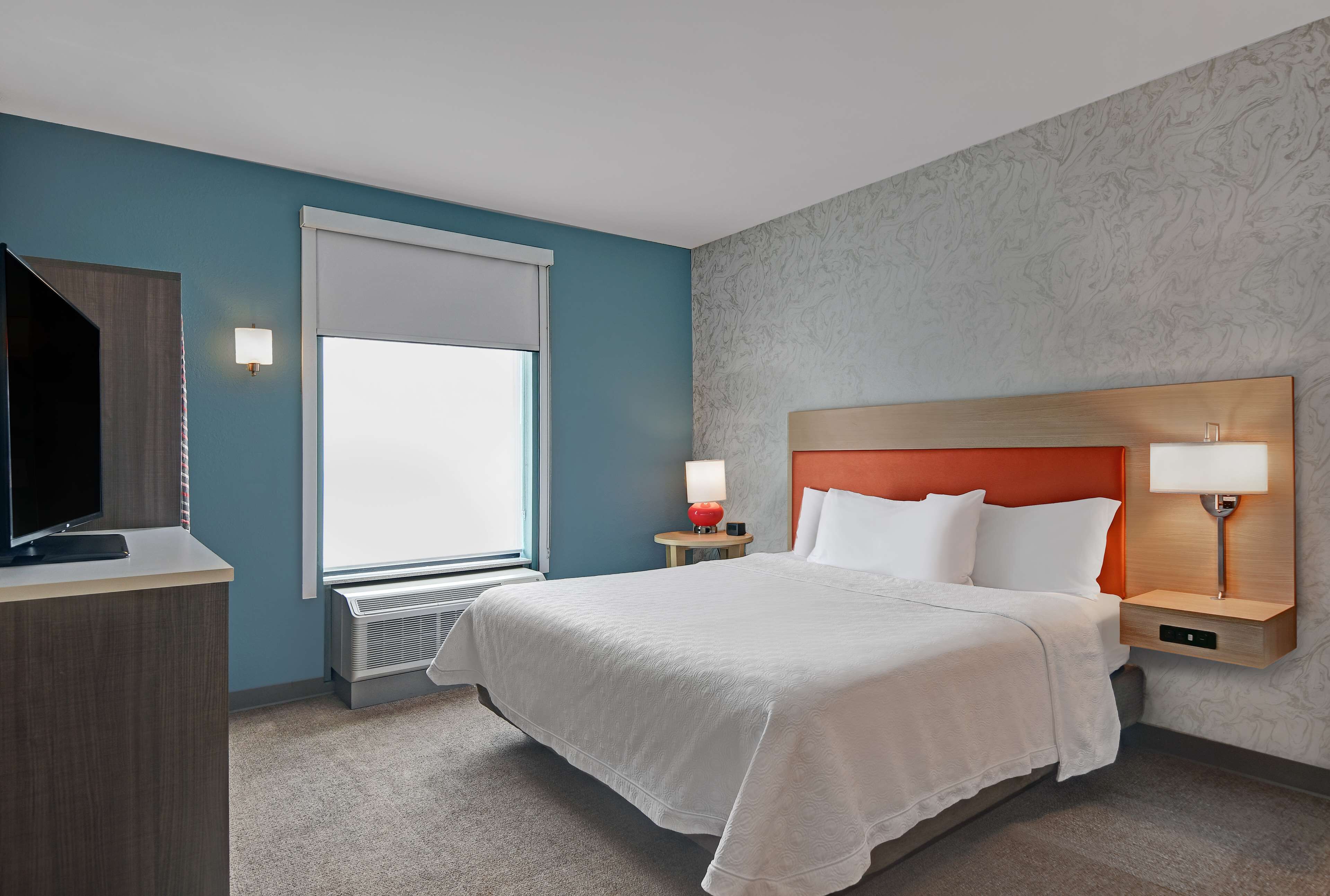 Habitación de hotel con cama, escritorio, televisor, ventana con persianas y pared con diseño decorativo.