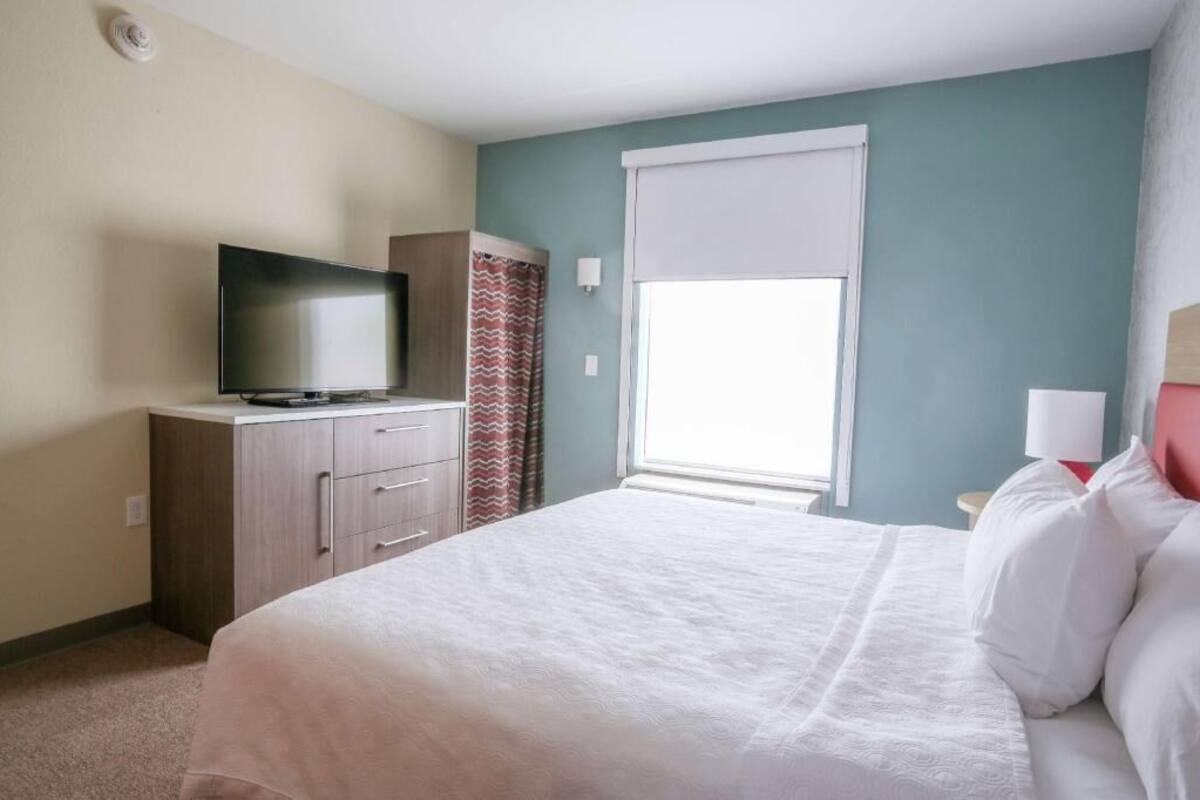 Ein Hotelzimmer mit Bett, Fernseher, Fenster mit Jalousie, Vorhang und Nachttisch mit Lampe.